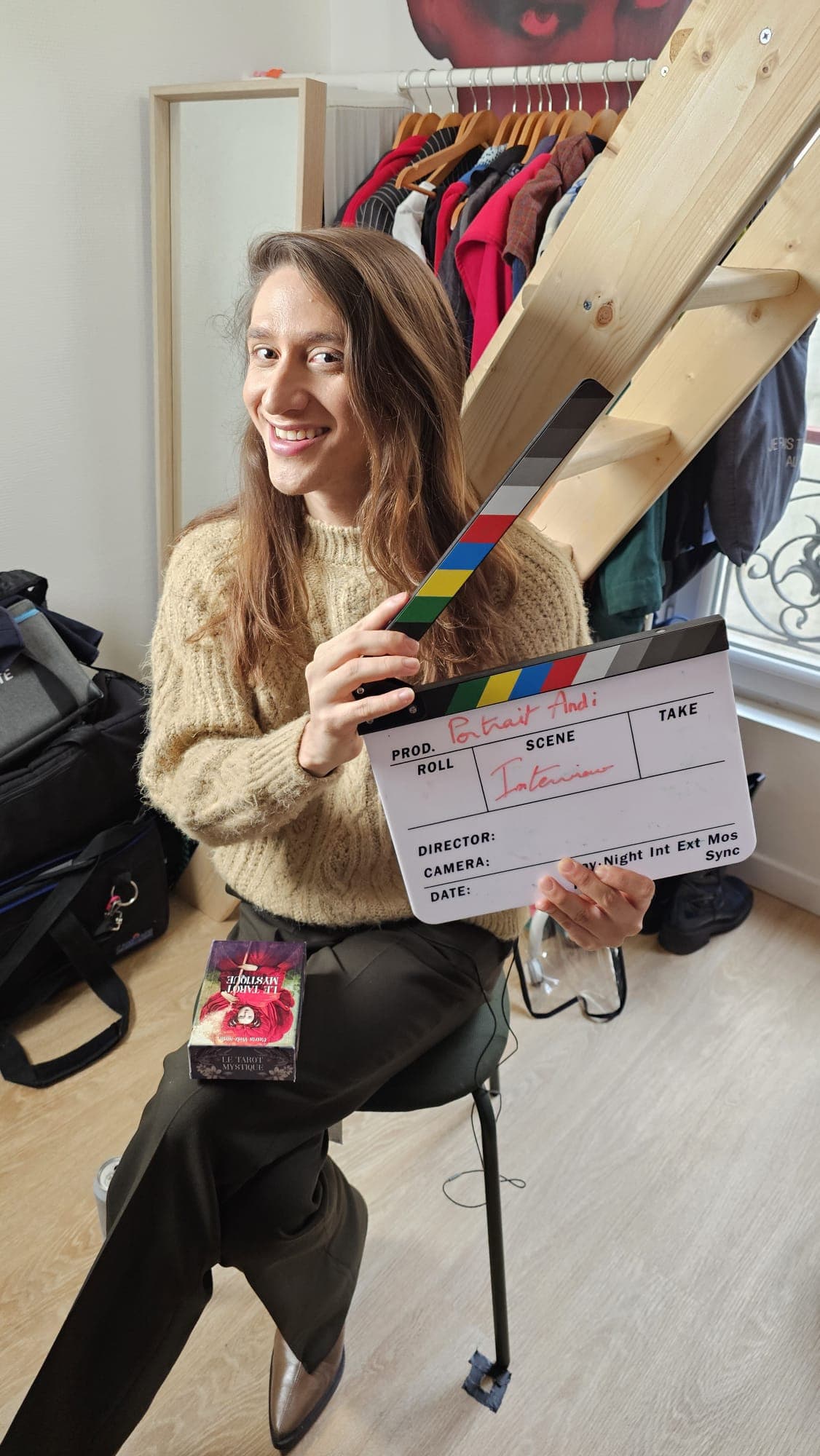 Andi sourit sur le tournage, un clap à la main marqué « Portrait Andi – Interview ».