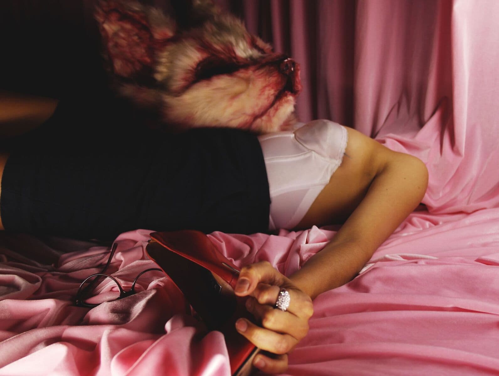 A performer in a fur animal mask lies on pink fabric, holding a red shoe, with glasses nearby.