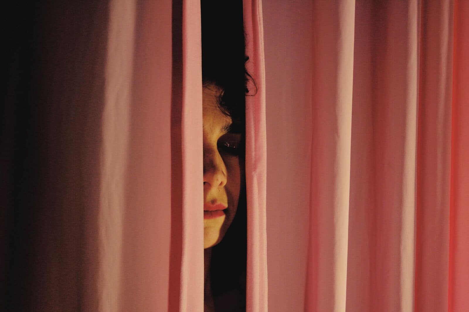 A face half hidden between pink curtains, in warm light.