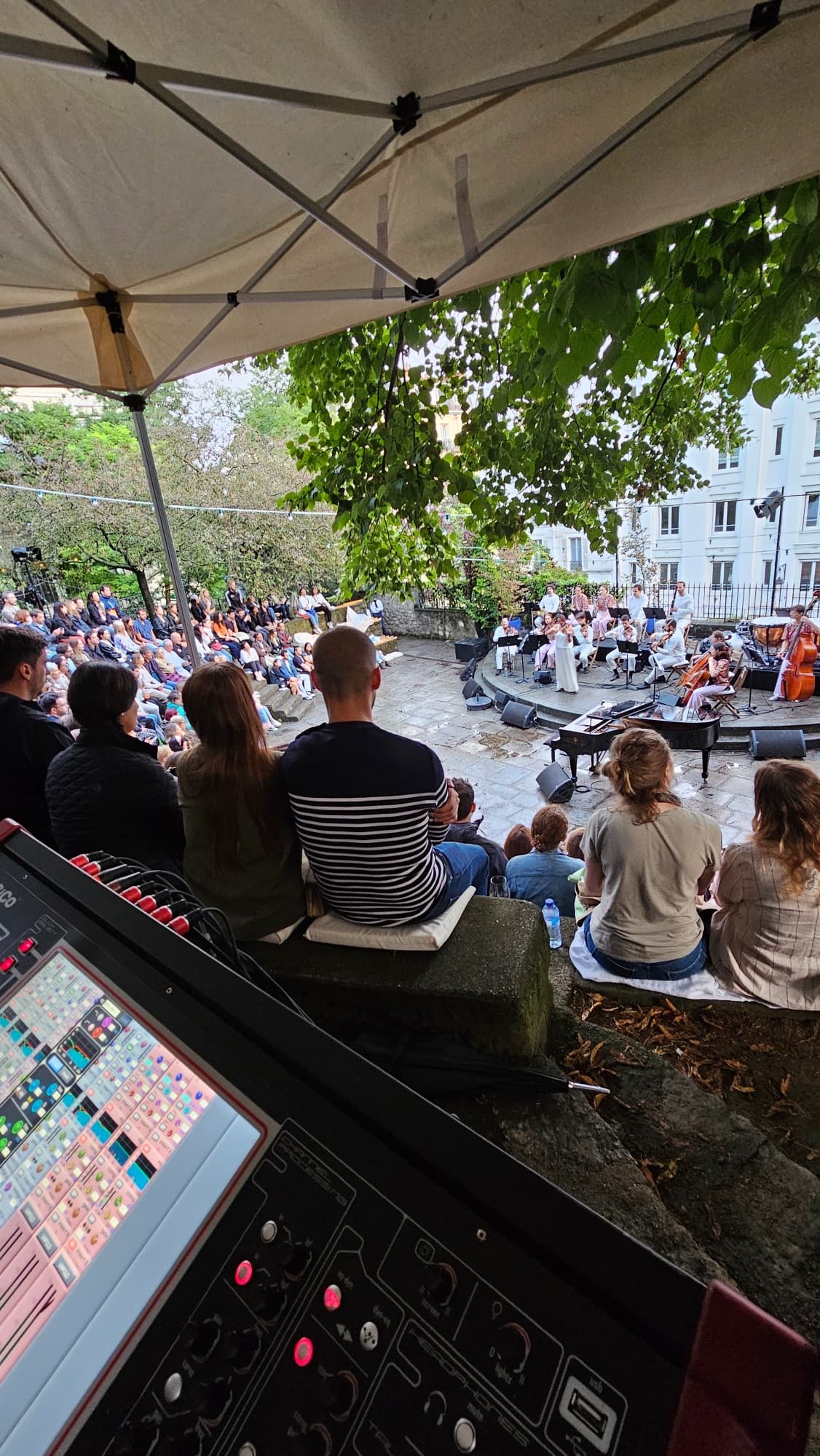 Vue depuis la régie, sous une tente : la console numérique au premier plan, le public sur les gradins de pierre et l’orchestre en blanc sur la scène ronde.