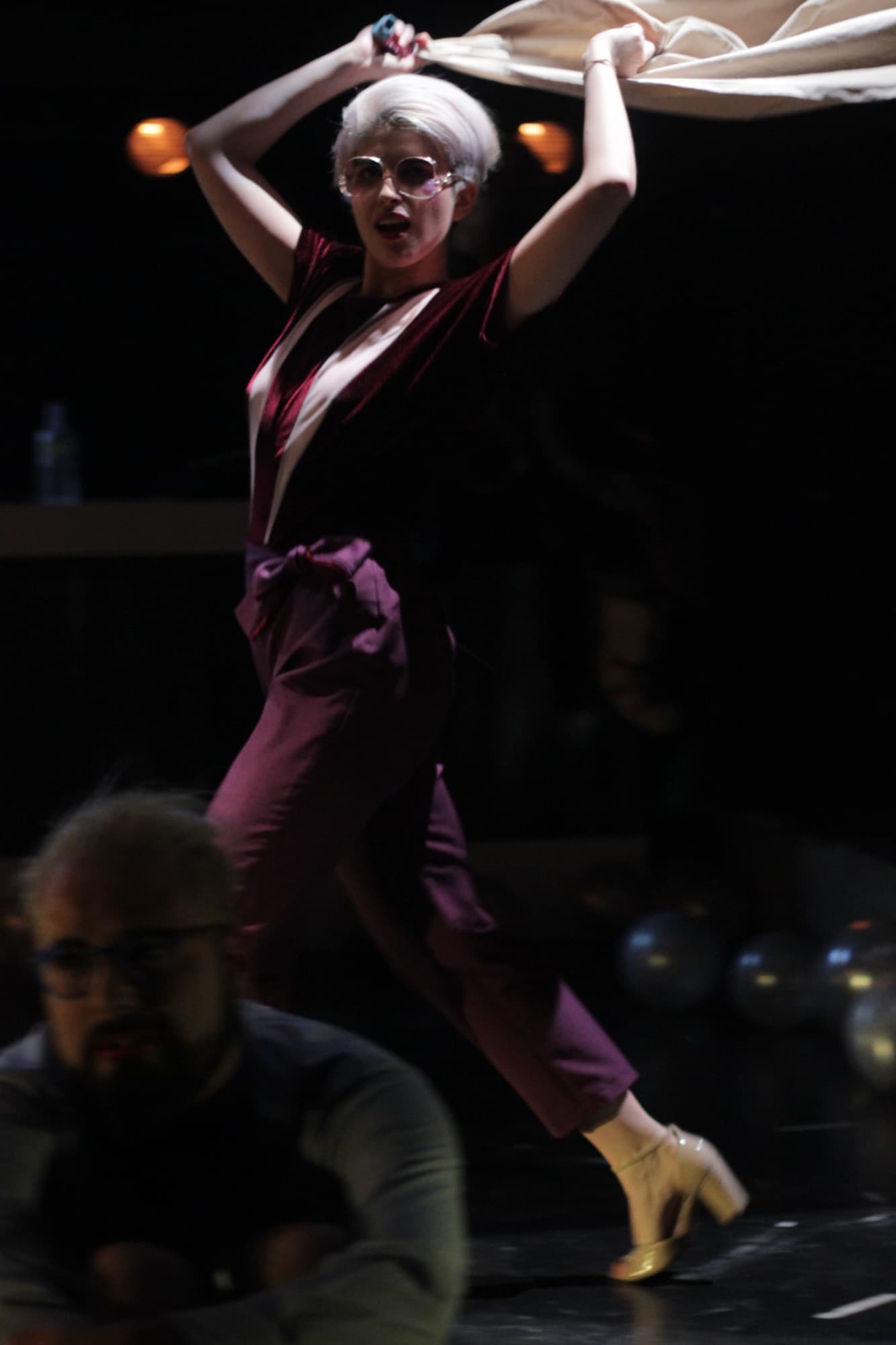 Gabriel as Glagolyev in Platonov, parading across the stage to show off the costume: white hair, large glasses, burgundy outfit, a sheet held overhead.