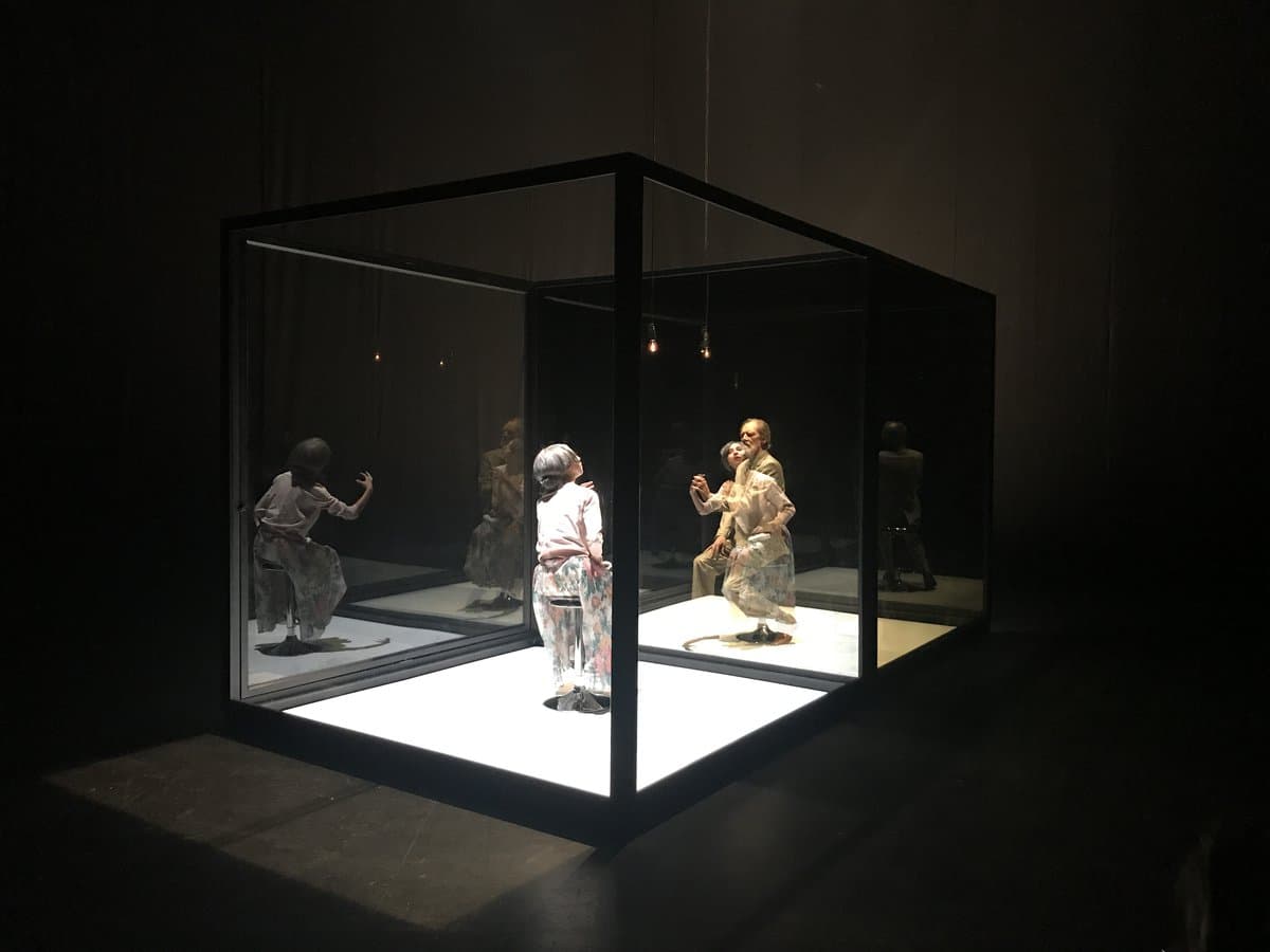 Locutorio: two performers inside a lit glass box on a dark stage, their reflections multiplied in the glass.