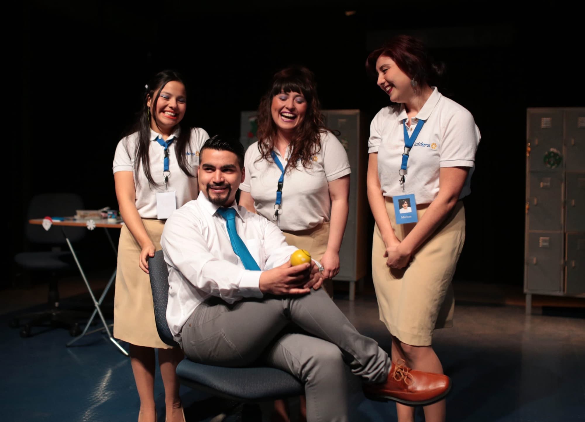 A man in a white shirt and blue tie sits holding a yellow ball, surrounded by three laughing cashiers.