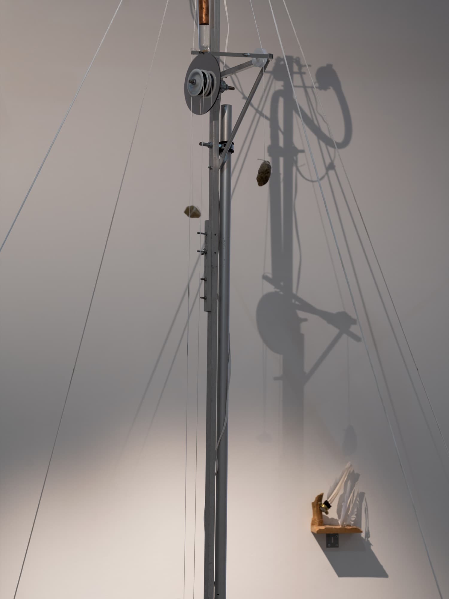 Détail d’une sculpture cinétique : un mât en aluminium avec une poulie et des pierres suspendues à des fils, son ombre sur le mur.