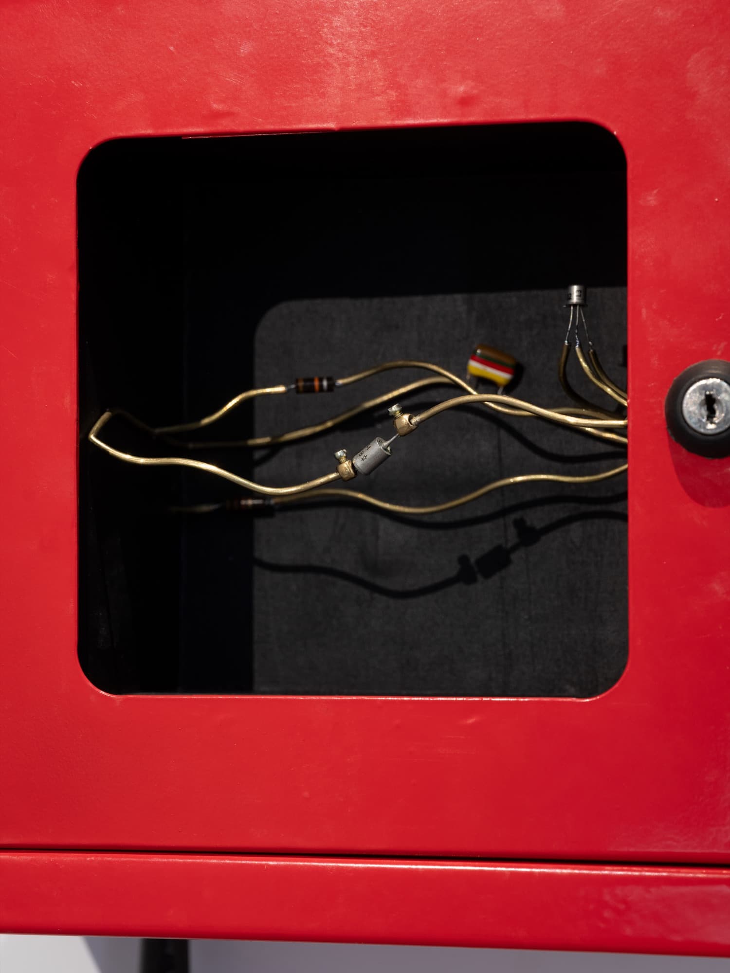 Gros plan sur un boîtier mural rouge : par sa fenêtre, des fils de laiton relient résistances, condensateur et transistor.