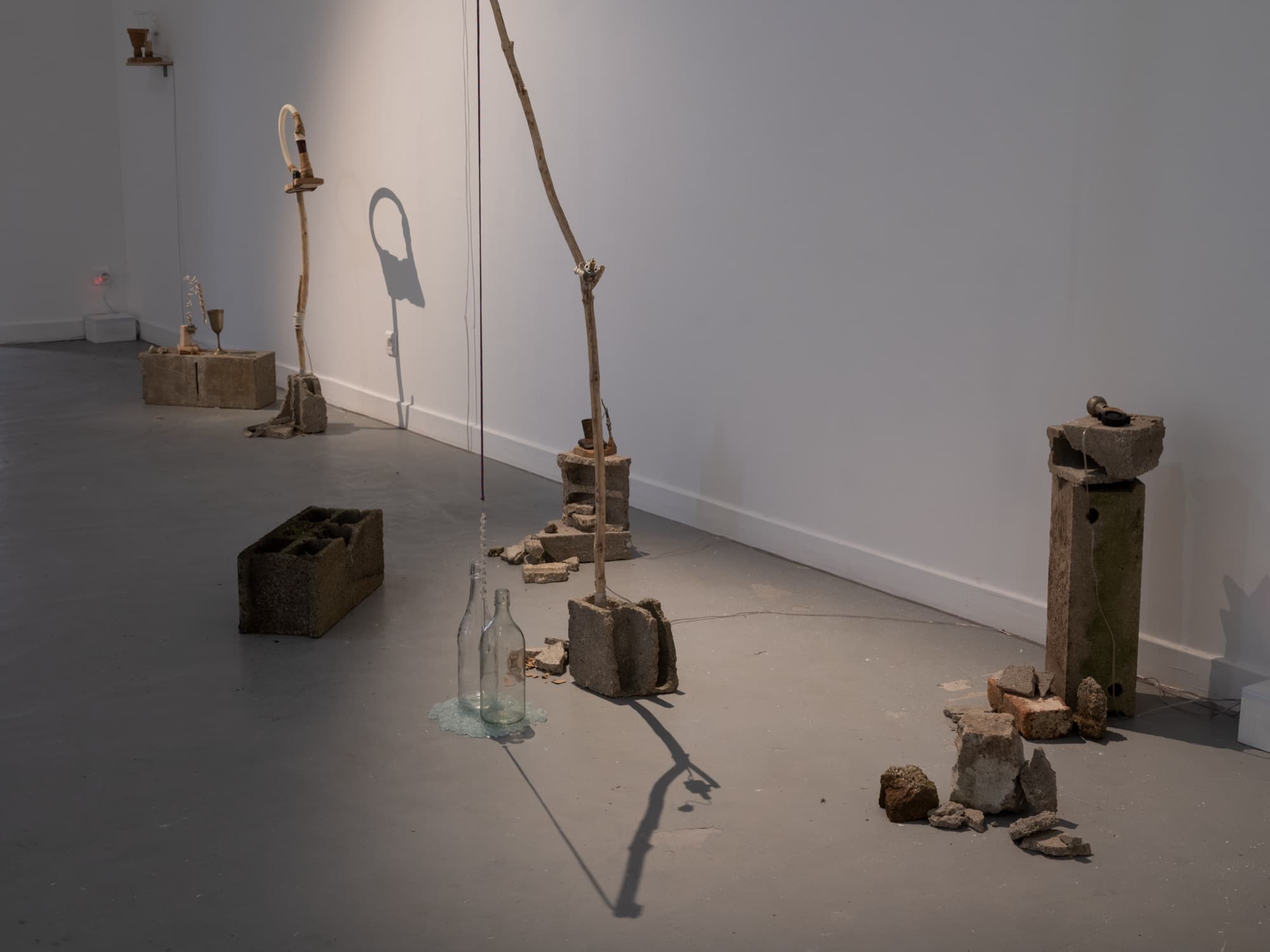 Une installation au sol de la galerie : parpaings, gravats, branches et deux bouteilles en verre sur une flaque de verre pilé.
