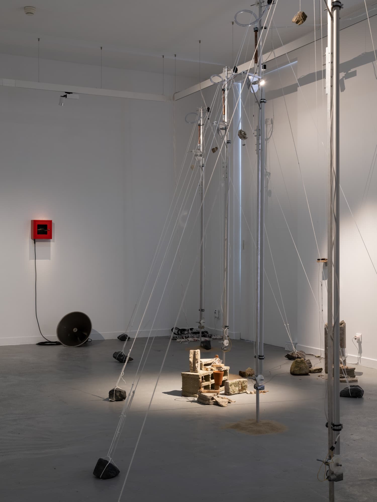 Vue d’ensemble de l’exposition : des mâts en aluminium tenus par des cordes blanches lestées de pierres, des gravats sous un projecteur, un boîtier mural rouge et un haut-parleur à pavillon au sol.